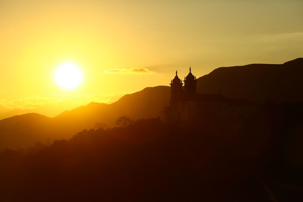 Silhueta de uma igreja em Ouro Preto durante o pôr do sol com o sol brilhando ao fundo sobre as montanhas